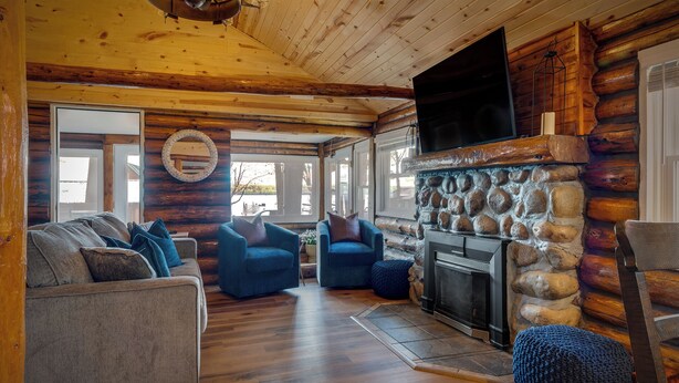 Interior de uma cabana de madeira com lareira de pedra, piso de madeira e uma TV grande fixada na parede.