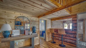 Interior rústico de cabana, com paredes e teto de madeira, área de refeições com mesa e cadeiras, e um espaço de estar acolhedor com um banco.
