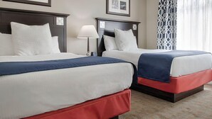 Dos camas matrimoniales con sábanas blancas y colchas rojas y azules, una mesita de noche con lámpara, y una cortina estampada.