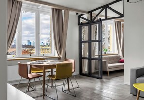A modern living room with a dining area, large windows with curtains, a sliding glass door, and a view of the cityscape. - Royal Baths Superior Apartment (Warszawa)