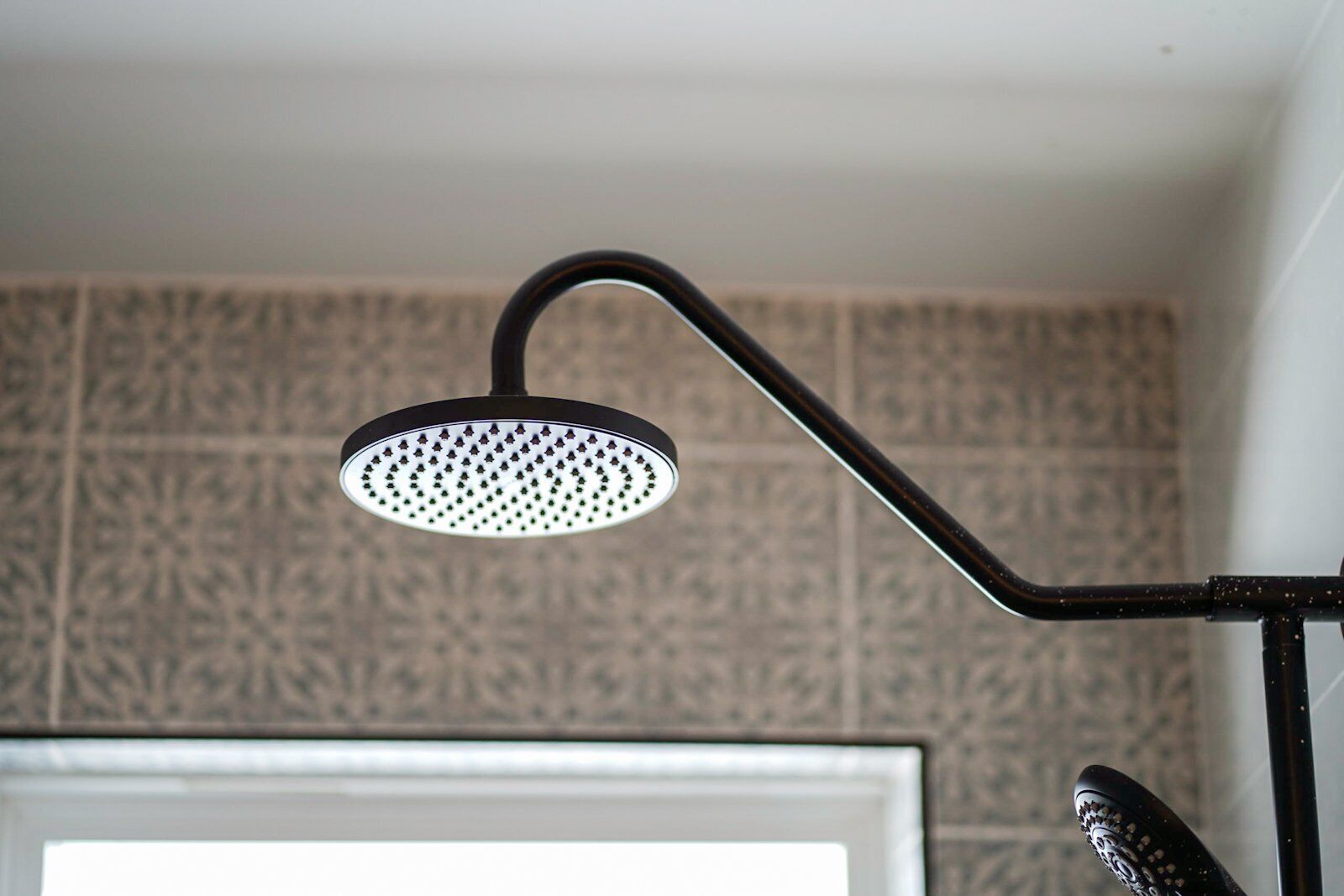 Un pommeau de douche doté d’un bras courbé, fixé sur un mur recouvert de carreaux décoratifs.