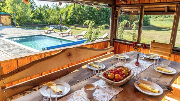 Mesa de comedor de madera dispuesta con platos, vasos y frutero, con vista a una piscina y una zona de estar al aire libre.