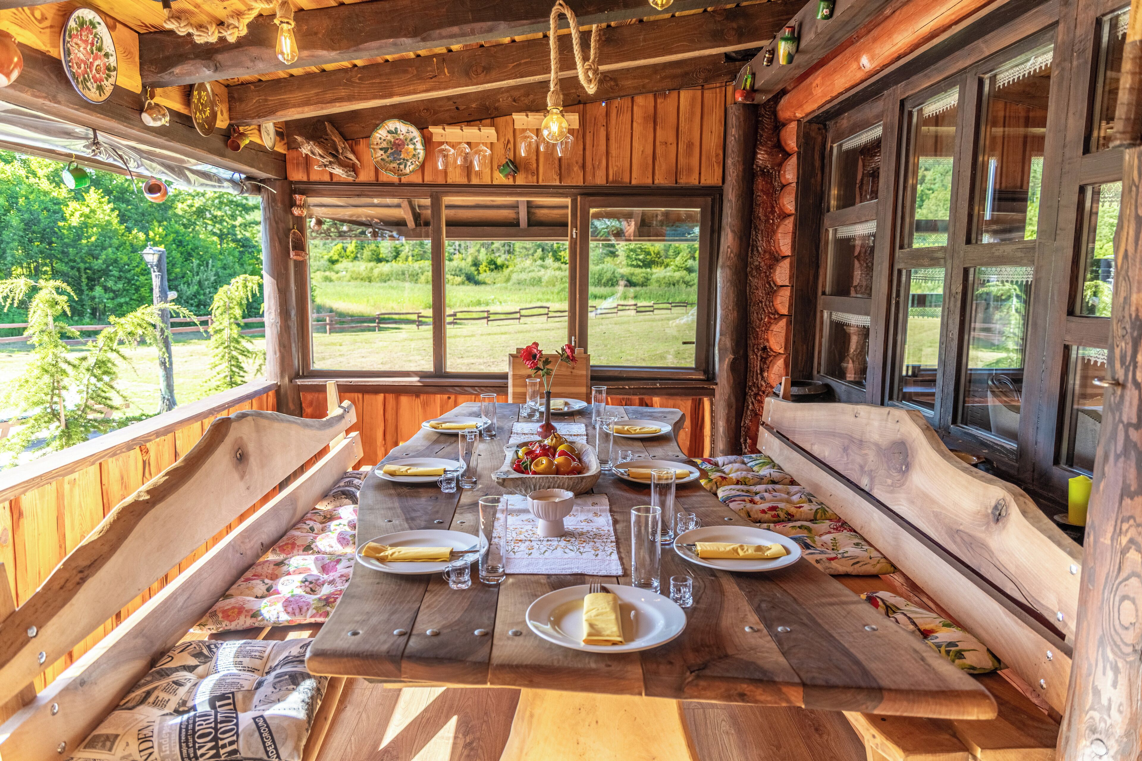 A rustic wooden dining area with a long table set for a meal, large windows offering a view of greenery, and a cozy interior with wooden beams and hanging light fixtures.