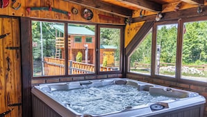 Un jacuzzi en una cabaña de madera con ventanas amplias y vistas a una terraza de madera y árboles.