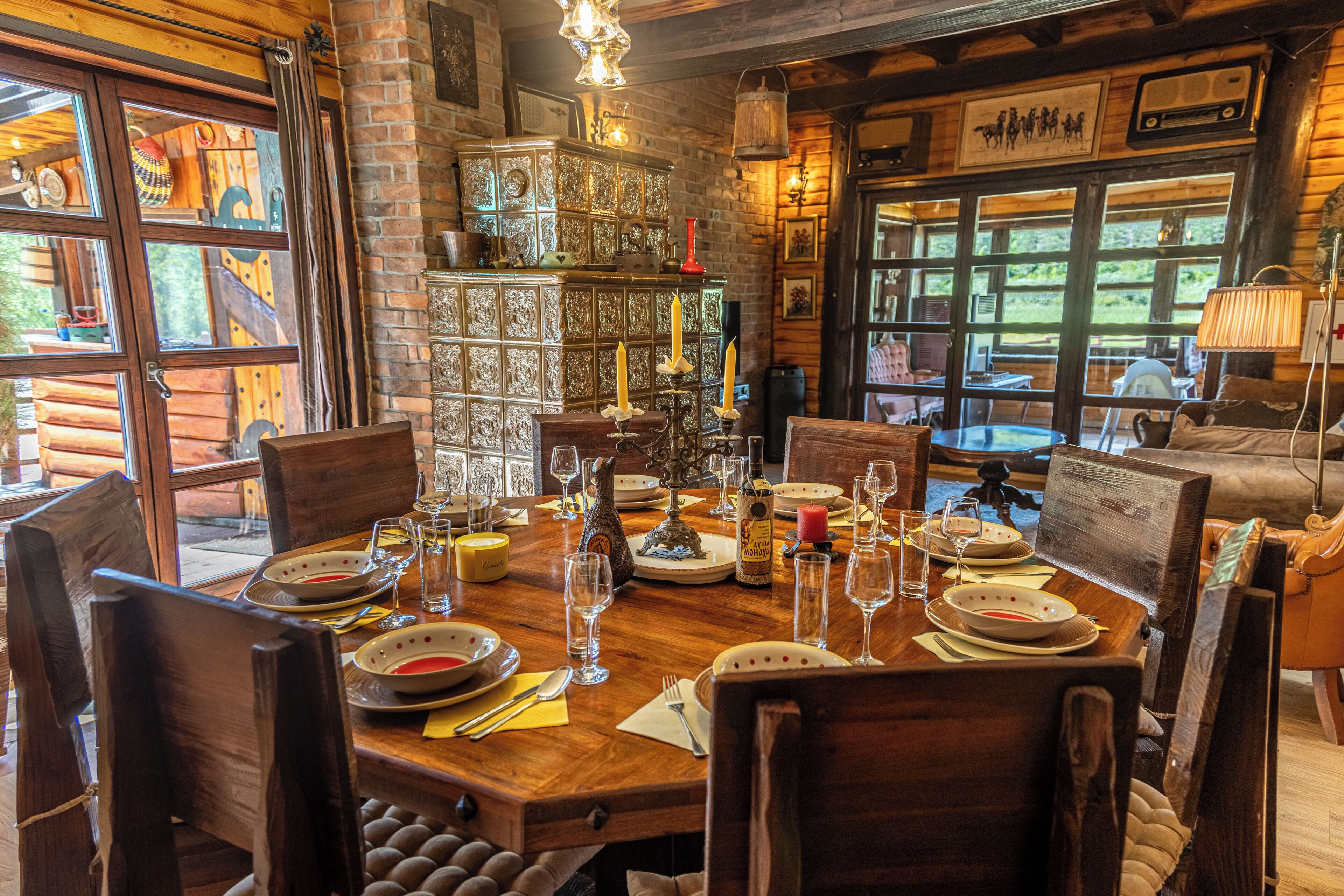 A wooden dining table set with plates, glasses, and candles, surrounded by chairs in a rustic room with brick walls and large windows.