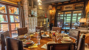Una mesa de comedor de madera dispuesta con platos, vasos y velas, rodeada de sillas en una habitación rústica con paredes de ladrillo y ventanas amplias.