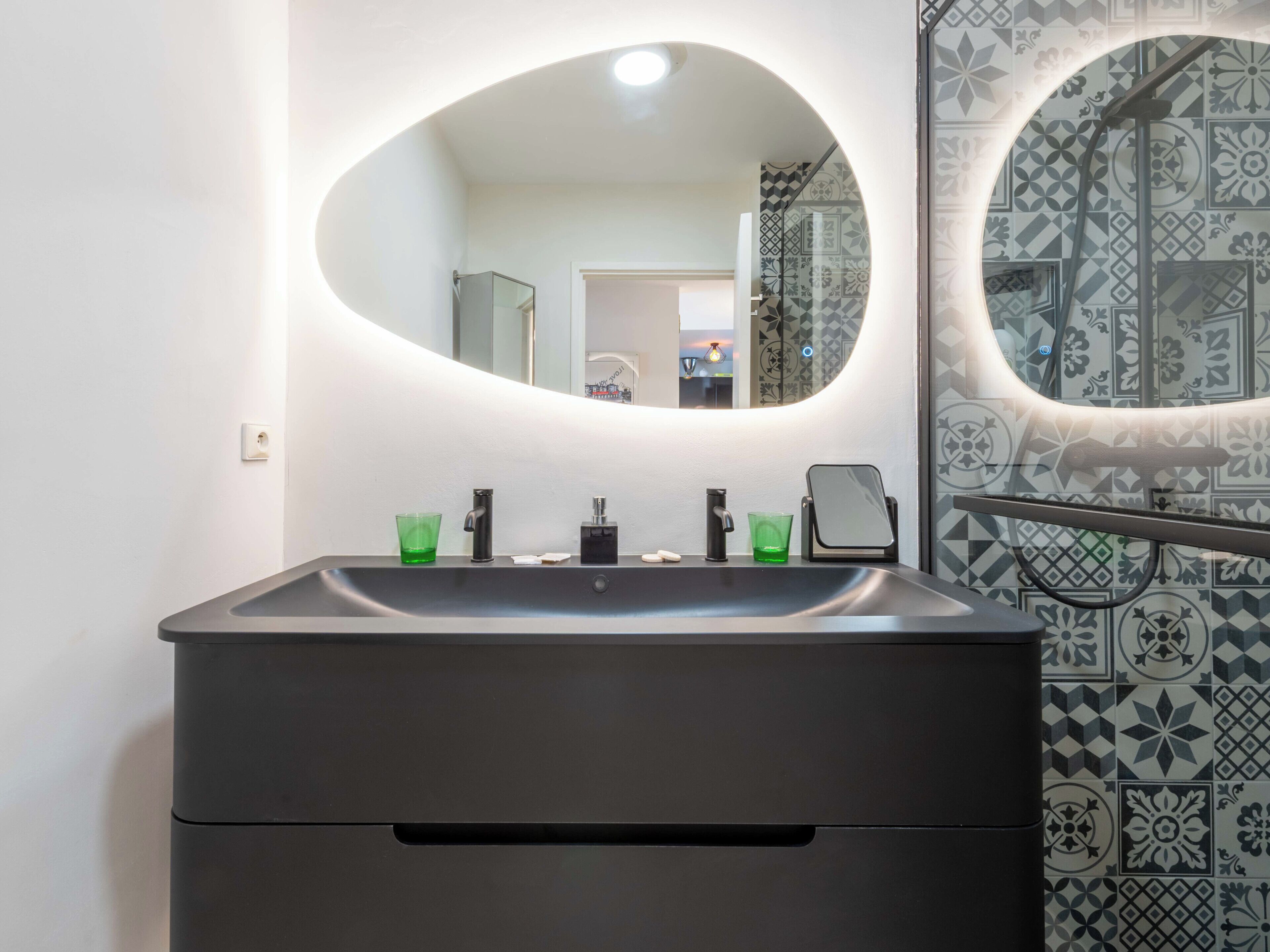 A modern bathroom with a large oval mirror, a black sink, and a patterned tile backsplash.