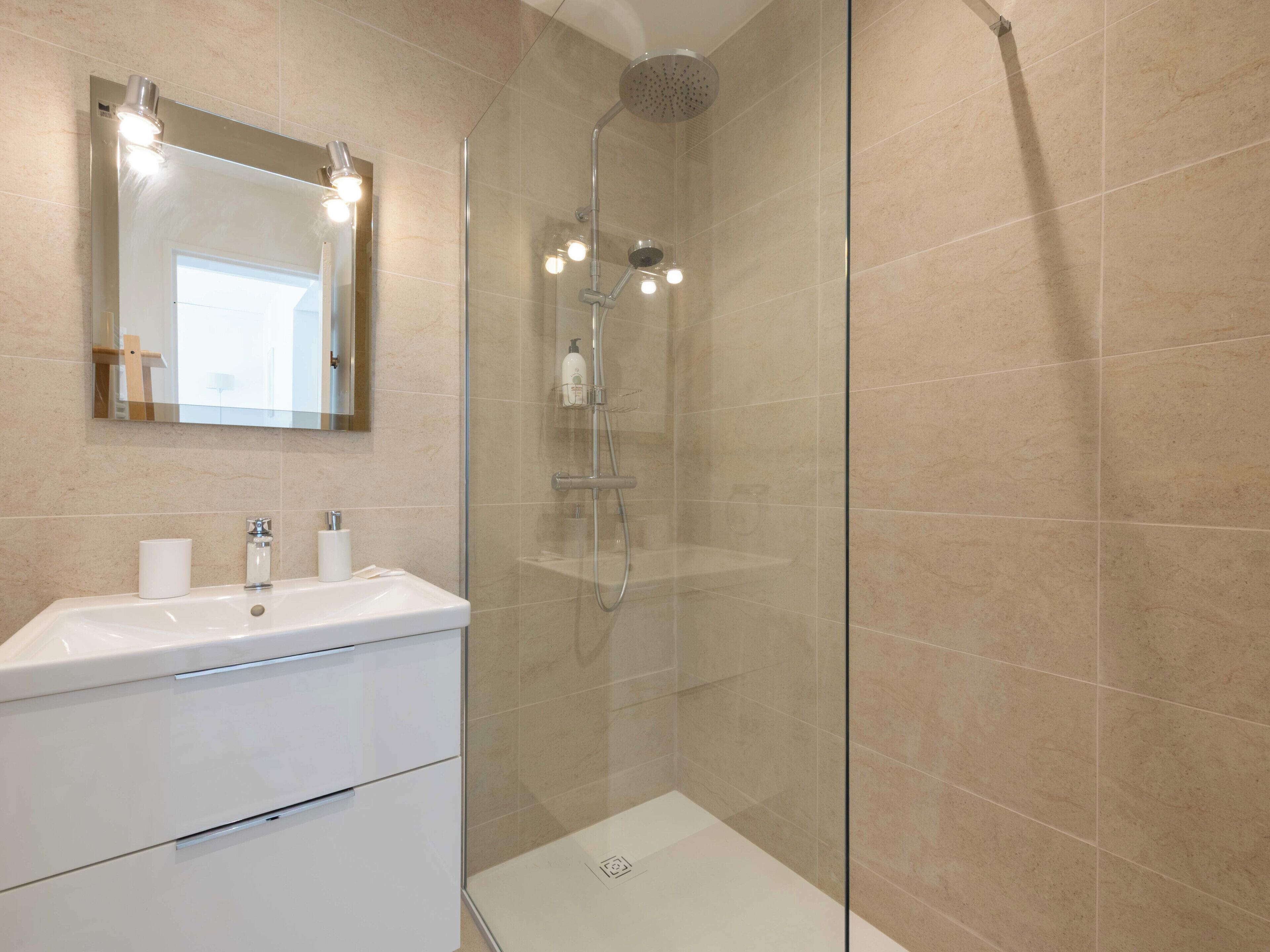 A modern bathroom with a glass-enclosed shower, a white sink with a mirror above it, and beige tiled walls.