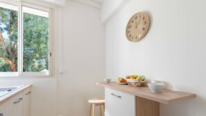 Una cocina con ventana, un reloj de madera, una cesta de frutas y un taburete.