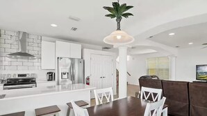 A modern kitchen with white cabinetry, a central island with stools, a dining table with chairs, and a hanging plant. - 4-Bedroom Villa in Flagler Beach with priority beach access (Flagler Beach)