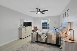 A bedroom with a bed, a ceiling fan, a dresser, a chair, a window, and artwork on the wall.
