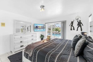 A modern bedroom with a bed, dresser, TV, and a view of a pool. - WATERFRONT - Walk to Beach, Pool/Spa, Kayaks, Bikes, Direct Gulf Access - Retro Retreat - Roelens (Cape Coral)