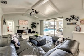 A modern living room with a black leather sofa, a coffee table, and a ceiling fan. There is a large window with a view of a swimming pool. - WATERFRONT - Walk to Beach, Pool/Spa, Kayaks, Bikes, Direct Gulf Access - Retro Retreat - Roelens (Cape Coral)
