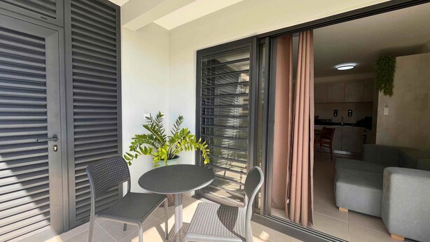 Un balcón con una mesa, dos sillas, una planta en maceta y un ventanal corredero que da a un espacio habitable con un sofá y una cocina al fondo.