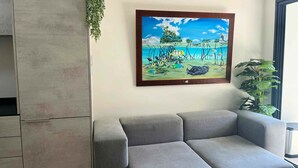 Una sala de estar con un sofá gris, un cuadro de una escena tropical y una planta.