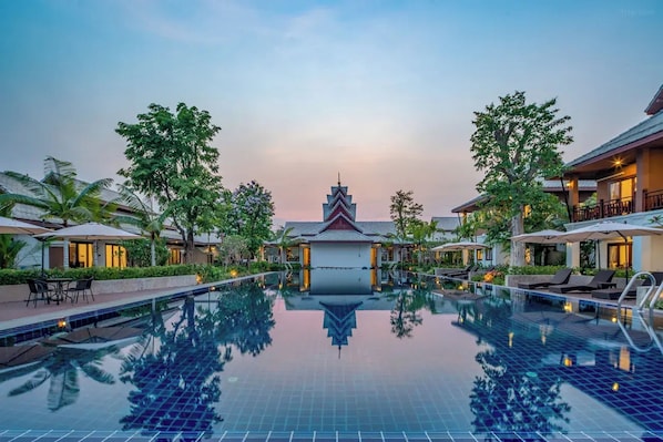 A swimming pool with a reflection of a building and trees, surrounded by lounge chairs and umbrellas.