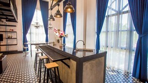 A modern kitchen with a central island, bar stools, pendant lighting, and a view through large windows with blue curtains. - Maerim Creek Resort Chiangmai (Mae Rim)