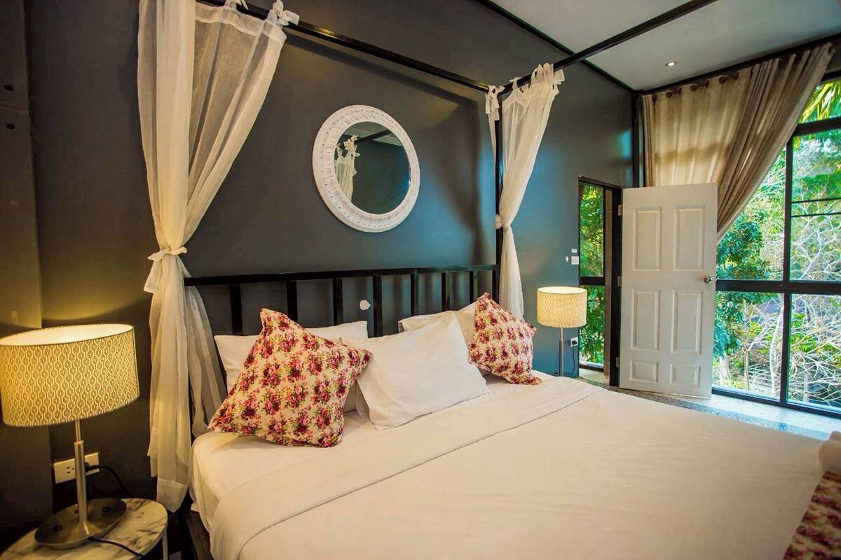 A bed with white bedding and floral pillows, a bedside lamp, a round mirror on the wall, and a view of greenery through a window.