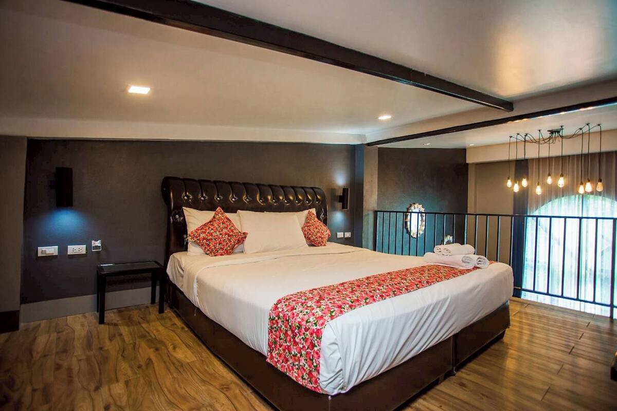 A modern hotel room with a large bed, a small bedside table, a dark headboard, and a balcony with a railing and hanging lights.