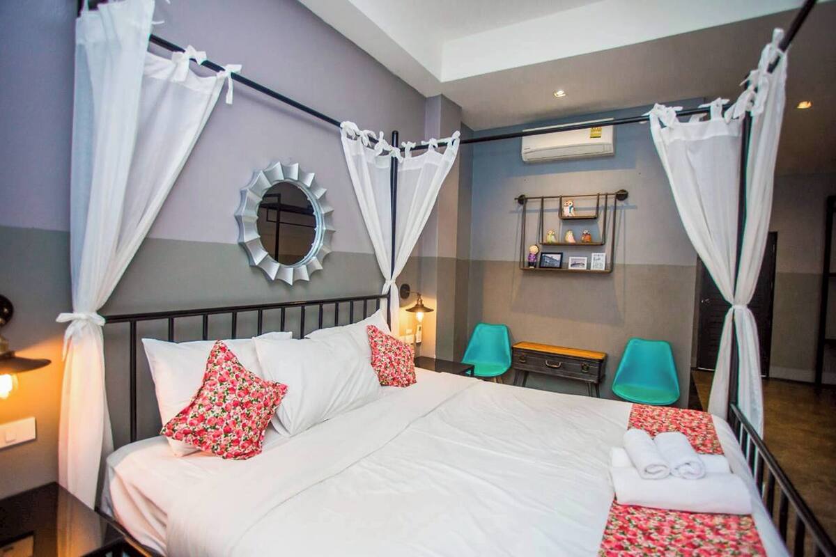 A neatly made bed with white and red floral pillows, a bedside table with a lamp, and a mirror on the wall.