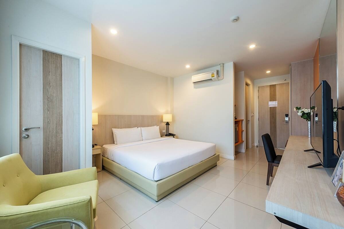 A hotel room with a bed, a desk, a chair, a TV, and an air conditioning unit.