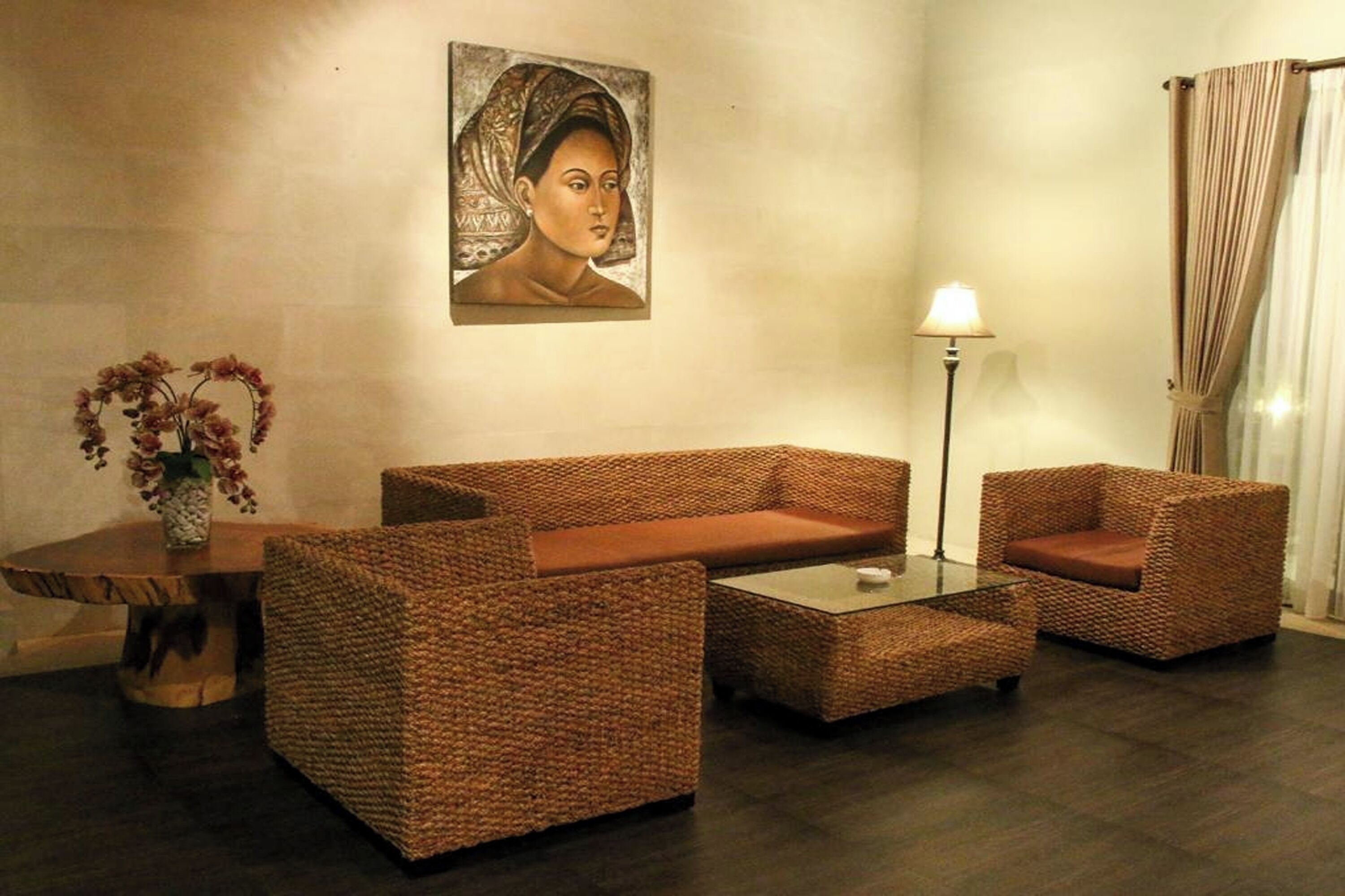A room with a woven sofa set, a small table with a vase of flowers, a floor lamp, and a painting on the wall.