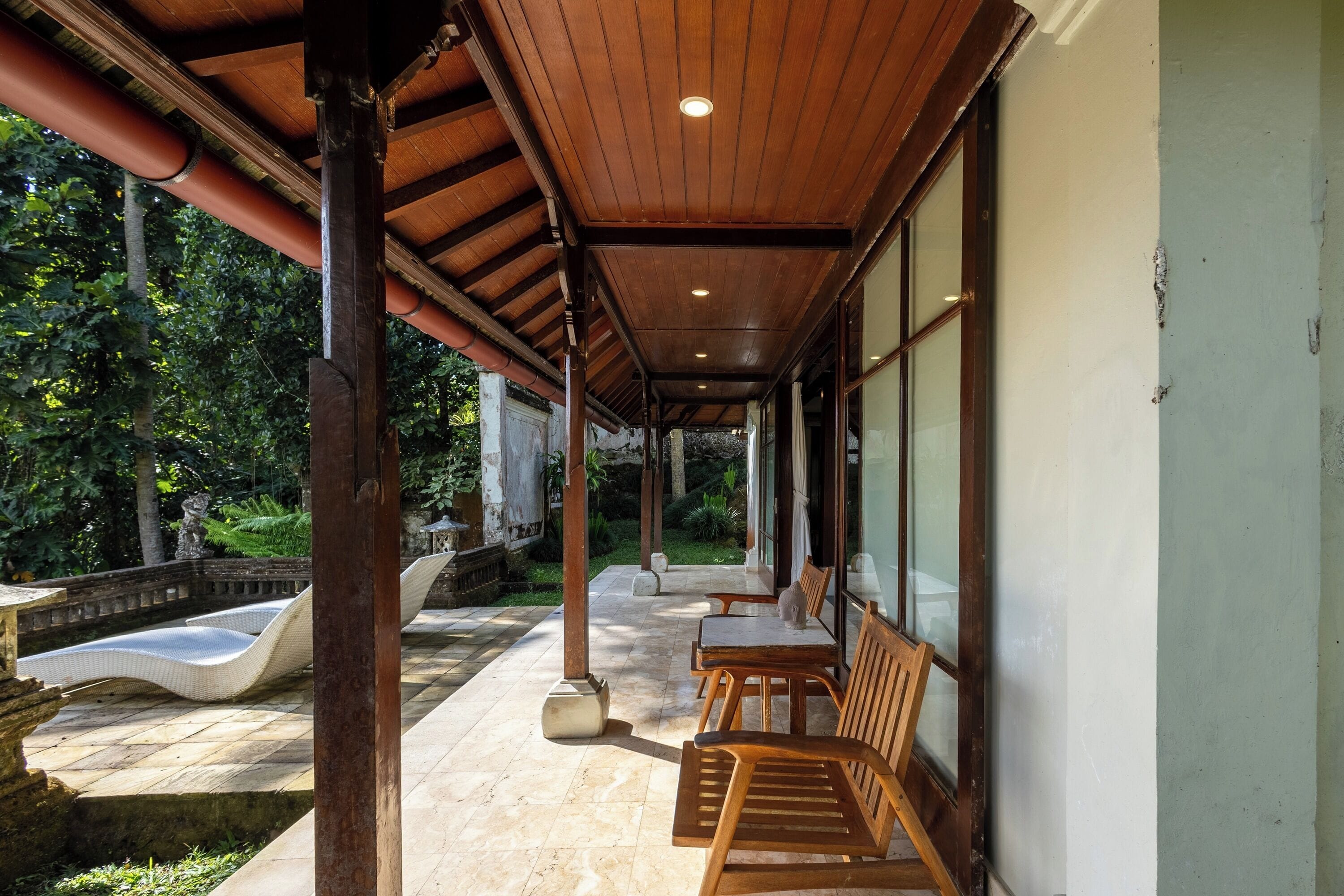 A covered outdoor seating area with wooden furniture and a stone pathway leading to a water feature.