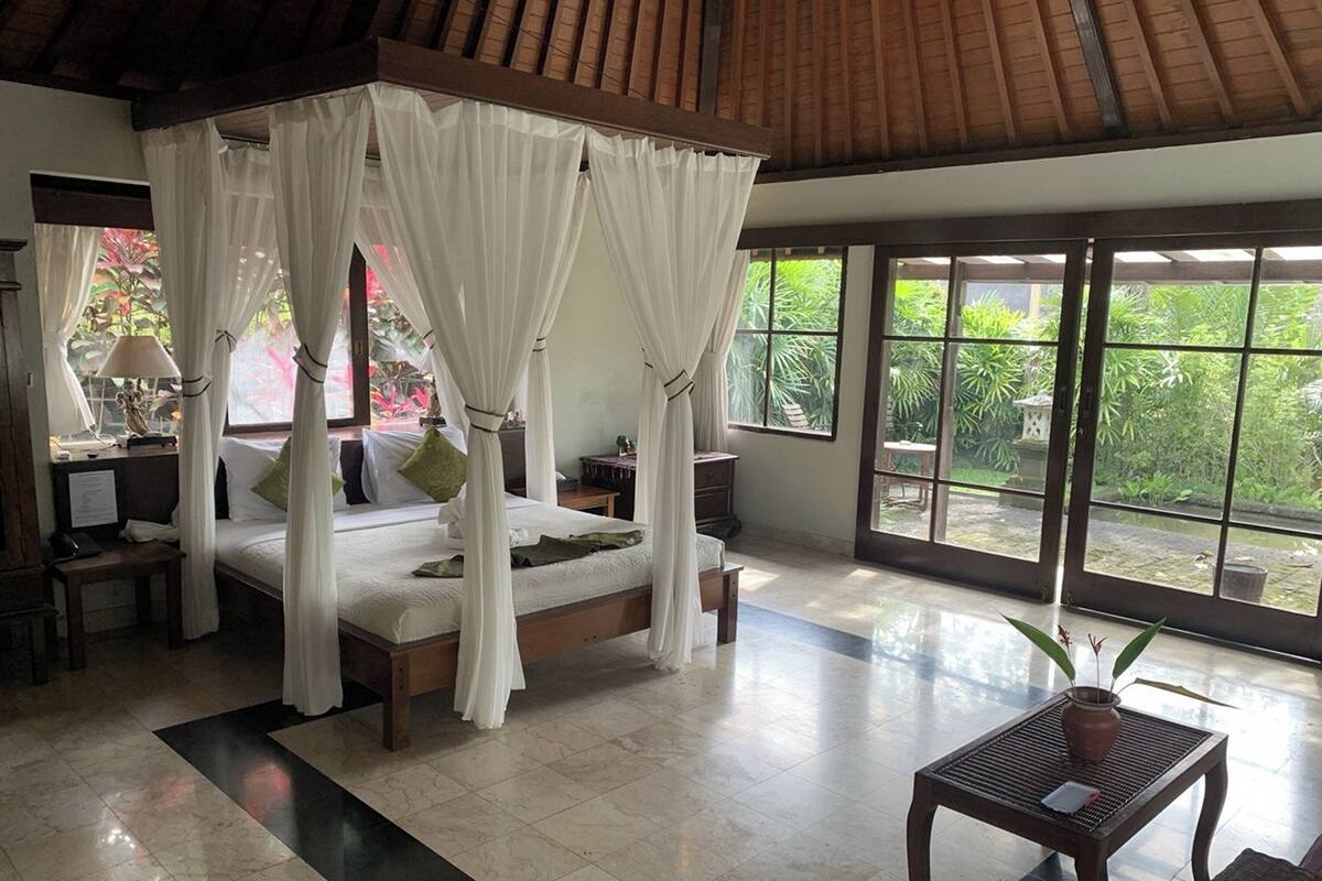 A four-poster bed with white drapes, a wooden nightstand, a small table with a plant, and a large window with a view of greenery.
