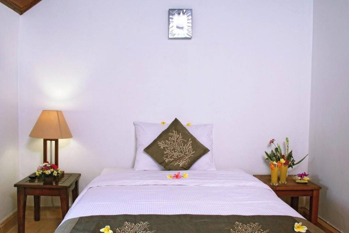 A neatly made bed with a decorative pillow, a bedside table with a lamp and flowers, and a wall clock.