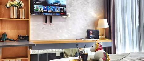 A modern hotel room with a large bed, a desk with a laptop, a TV mounted on the wall, and a plush unicorn toy on the bed.