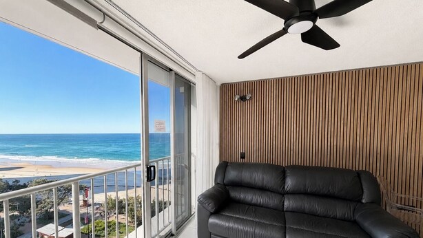A balcony with a view of the beach, a black leather sofa, and a ceiling fan. - Beachside Ocean View 1BR Apartment Central Surfers (Surfers Paradise)