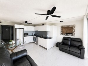 Una sala moderna con un sofá de cuero negro, una mesa de comedor con sillas, un área de cocina con armarios blancos y encimeras negras, y un ventilador de techo.