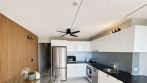 A modern kitchen with a glass dining table, stainless steel appliances, and a ceiling fan. - Beachside Ocean View 1BR Apartment Central Surfers (Surfers Paradise)