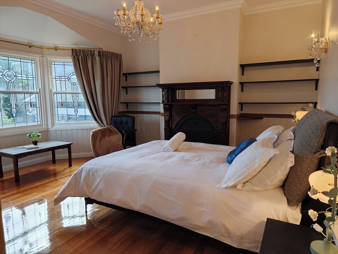 Ein geräumiges Schlafzimmer mit einem großen Bett, einem Holzkopfstück, einem Kamin, einer Chandelieber und Blick durch das Fenster.