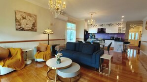 A spacious living room with a blue sofa, a round coffee table, and a chandelier. A kitchen is visible in the background with modern appliances. - 4BR Upper House Close to Blue Mountains Highlights (Katoomba)
