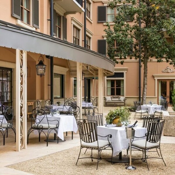 Un espace extérieur aménagé pour les repas, avec des tables et des chaises, une partie abritée et un bâtiment en arrière-plan.