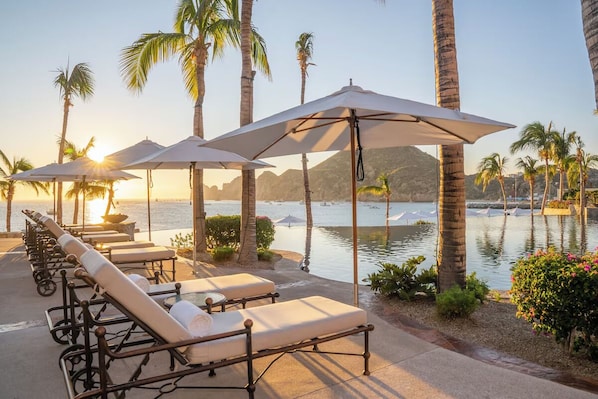 Un espace aménagé au bord de la piscine, avec des chaises longues et des parasols, des palmiers et une vue sur l’eau.