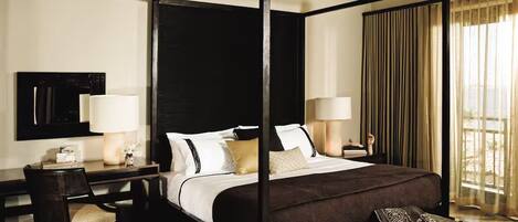 A four-poster bed with a dark headboard, a desk with a chair, a bedside table with lamps, and a patterned rug.