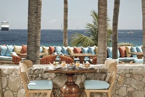 Un patio frente al mar con muros de piedra, sillas de madera y una mesa dispuesta para dos.