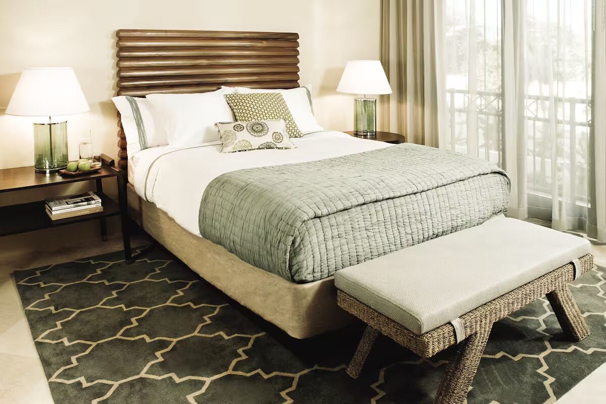 A neatly made bed with a green quilt, two bedside lamps, a nightstand with a book, and a patterned rug.