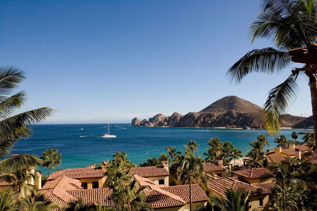 A coastal view with a clear sky, a mountain in the background, and a row of buildings with red-tiled roofs.