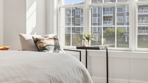 Et soveværelse med et stort vindue, en seng med hvid dyne, et natbord med en vase blomster, og udsigt til bygninger.