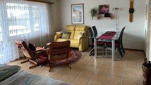A living room with a yellow sofa, a dining table with chairs, a wooden rocking chair, a patterned rug, and a ceiling fan. - Haus Montanara by Interhome (Leukerbad)