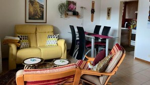 A living room with a yellow sofa, a dining table with chairs, and a kitchen area visible through an open doorway. - Haus Montanara by Interhome (Leukerbad)
