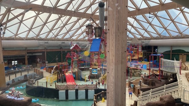 Parque acuático interior con techo de cristal amplio, diversas estructuras de juego y un tobogán de agua central.