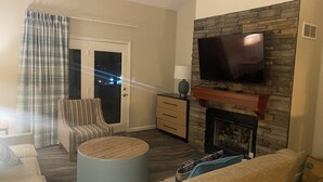 Una sala de estar con chimenea de piedra, televisor de pantalla plana, sofá, mesa de centro y una puerta con vistas al exterior.