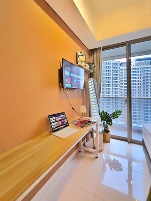 A modern hotel room with a flat-screen TV mounted on the wall, a laptop on a wooden desk, a bedside table with a lamp, and a large window offering a view of high-rise buildings. - Eldoria By SanLiving - Studio (Jakarta)