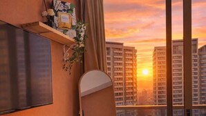 Ein Hotelzimmer mit großem Fenster, einem Flachbildfernseher, einem Schreibtisch mit Lampe, einem Sessel und einem kleinen Tisch mit Handtuch.