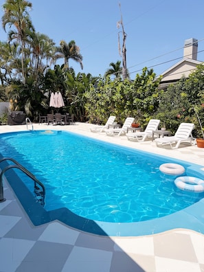 A swimming pool with lounge chairs and umbrellas, surrounded by tropical plants and a clear sky. - Poolside Palms & Oak; heated saltwater pool, 10 min to beach, A/C, dog-friendly (Venice)