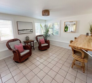 A spacious room with a wooden dining table, two wicker chairs with cushions, a small side table, and a large window with blinds. - Poolside Palms & Oak; heated saltwater pool, 10 min to beach, A/C, dog-friendly (Venice)